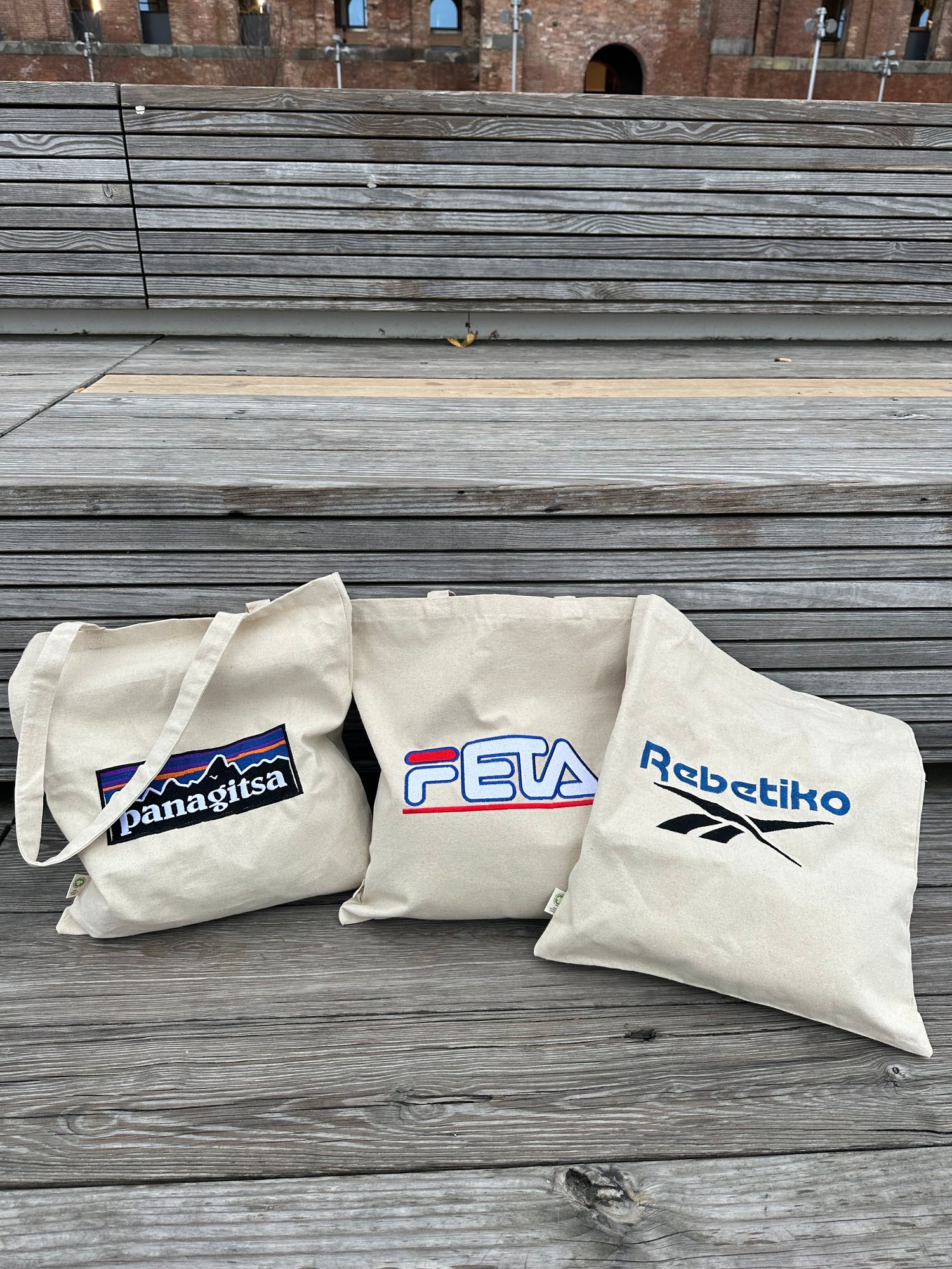 three bags laying on a bench with Rebetiko, Feta, and Panagitsa embroidered on them