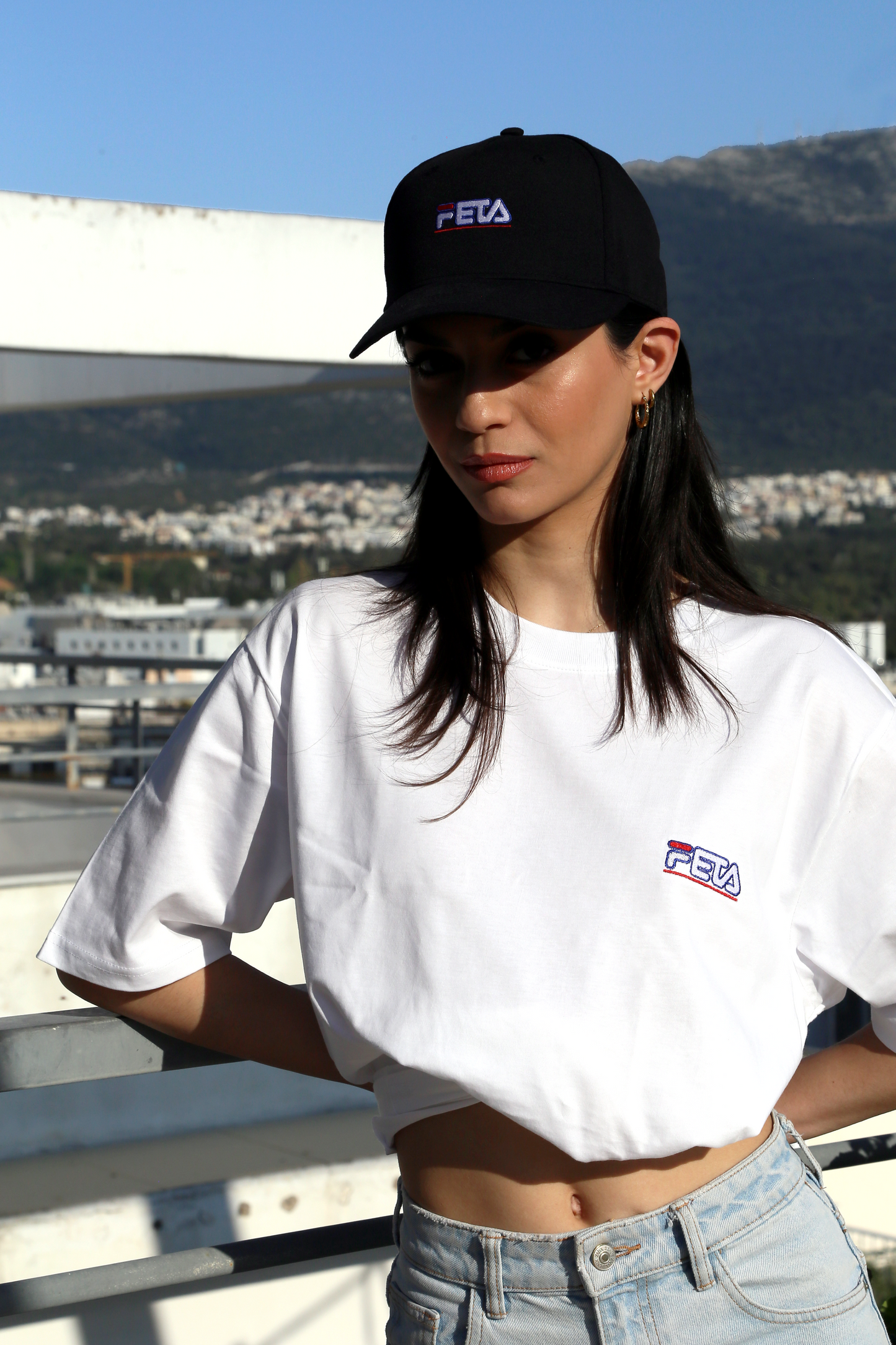 Woman wears black baseball hat with FETA logo inspired by FILA, white oversized t-shirt with Feta logo on the left hand side of the chest, inspired logo by FILA 